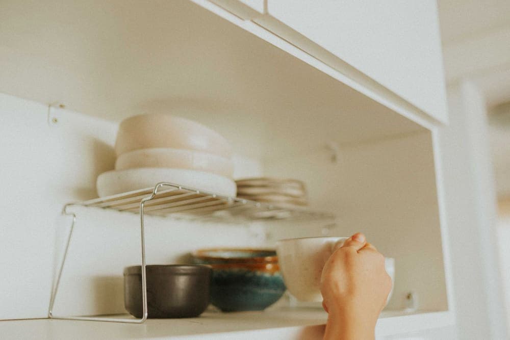A hand reaching for a mug in a kitchen cabinet fitted with a wire shelf riser