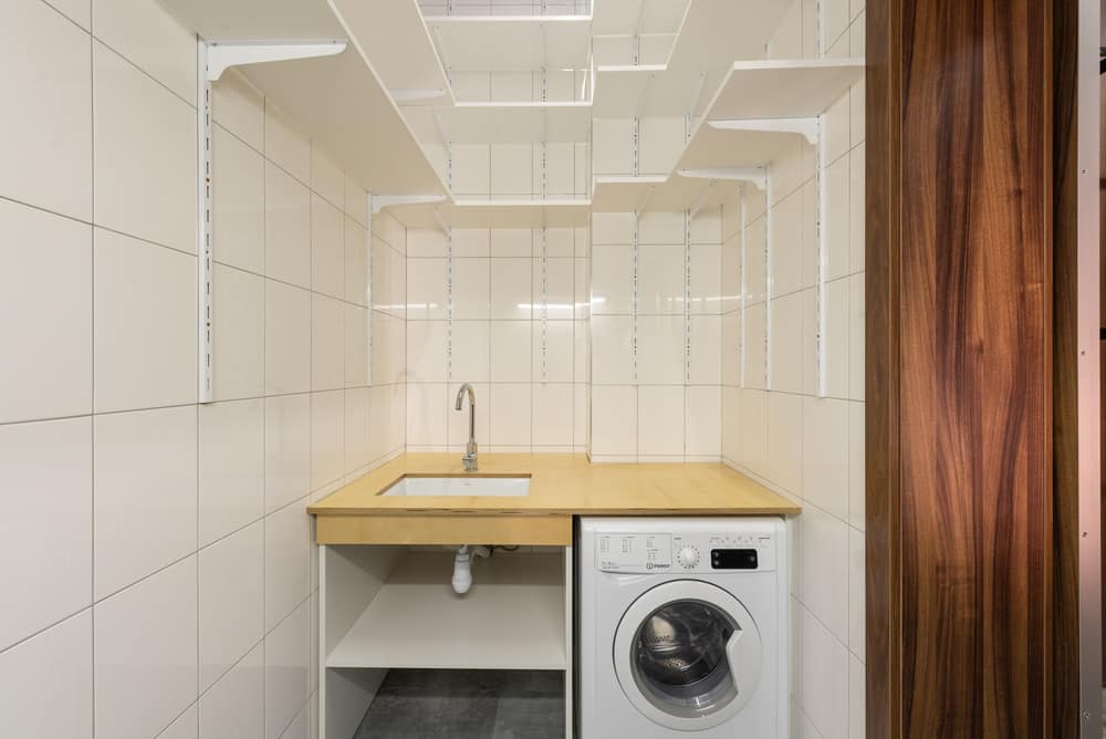 A washing machine under a wooden counter in a narrow tiled laundry room with empty wall shelves above